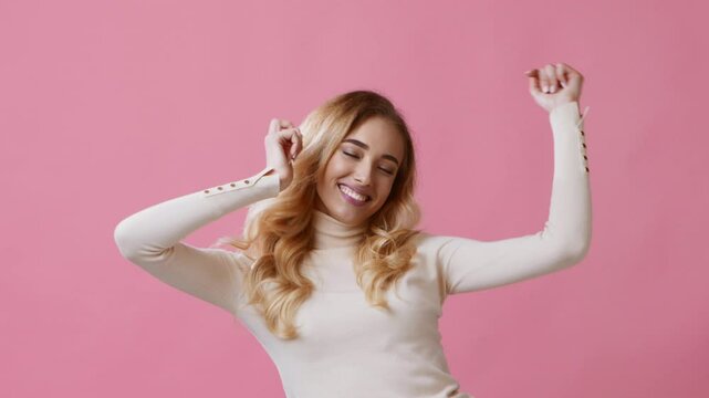 A young woman with wavy hair dances energetically, expressing joy and freedom. The vibrant pink background adds to the lively atmosphere, creating a cheerful mood.