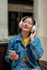 A relaxed woman is enjoying the music through her headphones while sitting on a bench in the city.