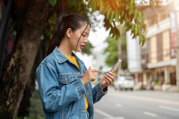 A side view of a woman standing on a sidewalk, checking her smartphone as she books a taxi on an app