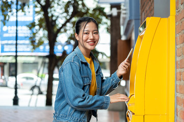 A beautiful Asian woman in a denim jacket is standing in front of an ATM, smiling at the camera.