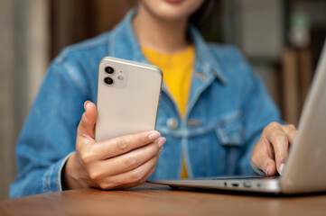 A woman holding her smartphone while working on her laptop, working remotely from a coffee shop.