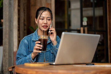 A woman is on the phone with her colleague while working on her laptop remotely from a coffee shop.