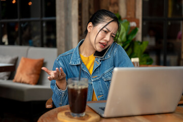 A stressed Asian woman is on the phone while working on her laptop remotely from a coffee shop.