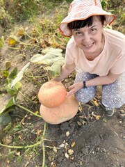 top view of a smiling female farmer picking pink pumpkins in a garden bed among green leaves on a garden plot in autumn, The concept of growing environmentally friendly products