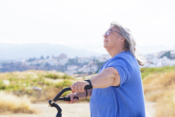 overweight white-haired woman riding a retro-style bicycle with a mountain landscape with a village in the background