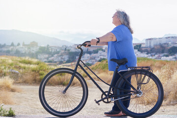 Obraz premium an overweight mature woman rides a black retro-style bicycle against a cloudy mountain backdrop with a village in the background