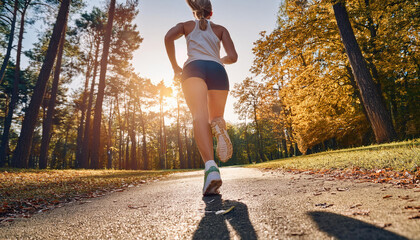 A runner woman running alone toward the sun in the park with tree on both side of the road. 