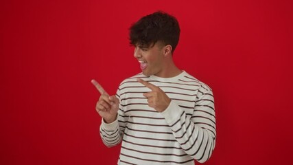 Man smiling confidently in stripes t-shirt, young hispanic male standing and pointing thumbs up to the back over isolated red background