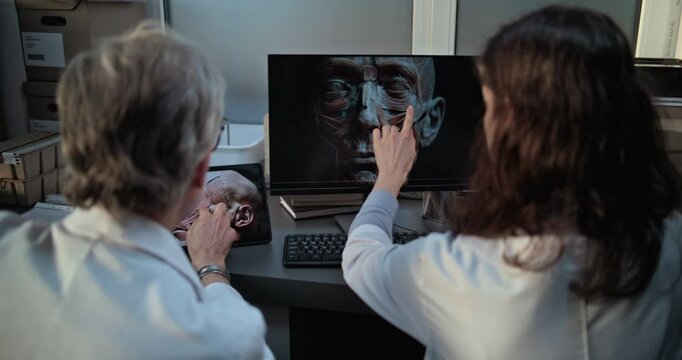 Two diverse scientists discuss 3D model of human head anatomy displayed on PC and tablet computer screens, conduct historical research. Archaeologists or paleontologists work in archaeological lab.