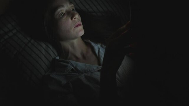 A young woman in bed uses her smartphone to surf the Internet at night. The light from the phone screen