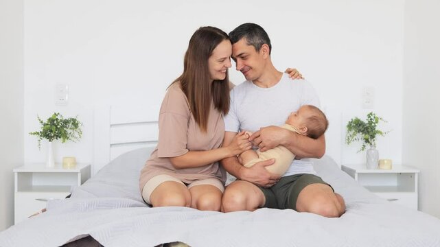 Mother and father hugging baby while sitting on bed parents cuddling their newborn and enjoying time together kissing in cozy intimate bedroom meeting new family members