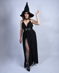 Full length portrait of beautiful female red head model wearing  glamorous black witch dress with pointy hat halloween costume.   walking pose with gestural arms, isolated figure on studio background