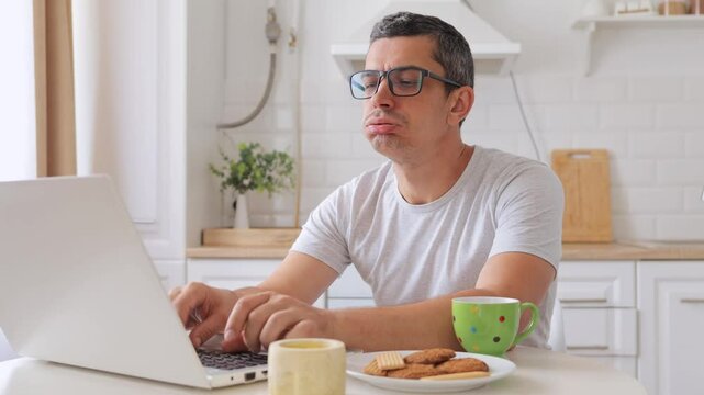 Despair man sitting at table in kitchen working on laptop angrily yelling at screen while browsing website frustrated by slow internet connection overwhelmed with remote job tasks