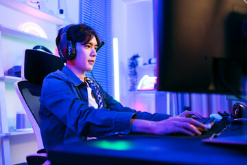 A young Asian man professional gamer wearing jeans jacket sits on a chair with a gaming table with pc, keyboard, monitor, microphone. Prepare for competition, cast gameplay or record a podcast. © artitwpd