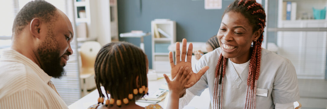 Doctor giving high-five to child during medical consultation as father watches proudly. Doctor smiling and showing support for child's achievement in healthcare setting