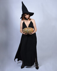 portrait of beautiful female model wearing glamorous black witch dress, pointy hat halloween costume. walking pose holding pumpkin, isolated figure on studio background