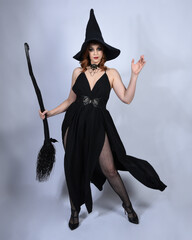 portrait of beautiful female model wearing glamorous black witch dress, pointy hat halloween costume. walking pose holding broomstick, isolated figure on studio background