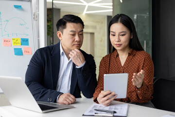 Asian business professionals engaging in collaborative discussion using tablet and laptop. Man and woman focused on productive teamwork and planning at modern office workspace.
