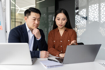 Asian business professionals working together on project in modern office setting. Two colleagues focused on laptop screen, discussing strategy and making important business decisions.