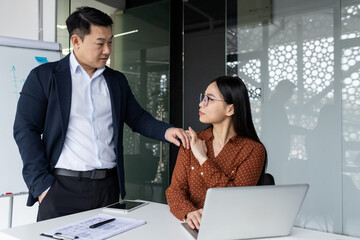 Office scene with male boss touching female subordinate's shoulder. Female employee looks...
