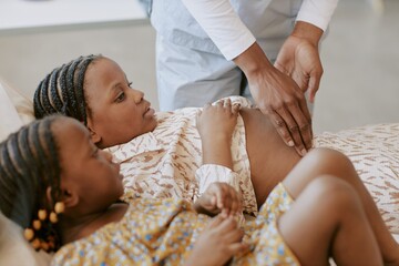 Medical professional examining an African childs abdomen on a bed while another child watches....