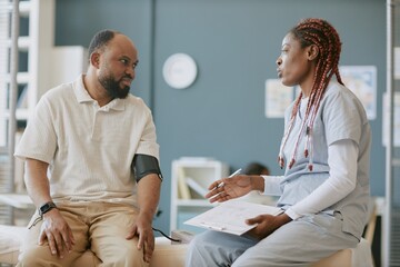 Obraz premium African American male patient sitting on hospital bed engaging with nurse while holding clipboard in medical office setup, displaying focused expressions and gestures