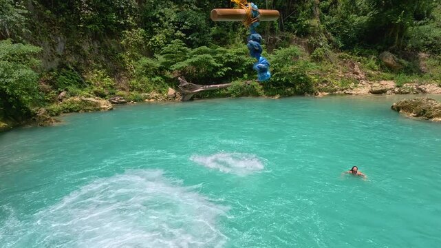 A man swings into bright blue water while canyoneering at Kawasan Falls in Cebu, Philippines. he jumps and swings from a platform and then falls into the water below where a woman is swimming.