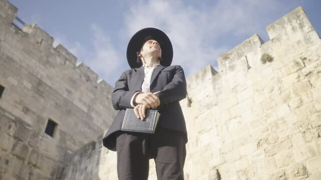 Jerusalem Old City, Israel, an orthodox Jewish man stands in front of the city walls wearing a black suit and hat and holding a prayer book on a sunny day. Stabilized low angle pan across the scene.