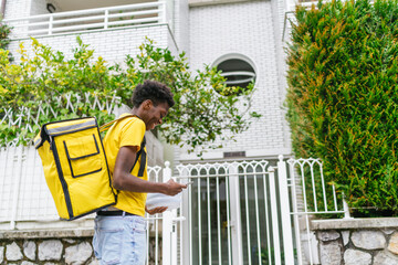 Smiling food delivery man with a yellow backpack, checking his phone in front of a house