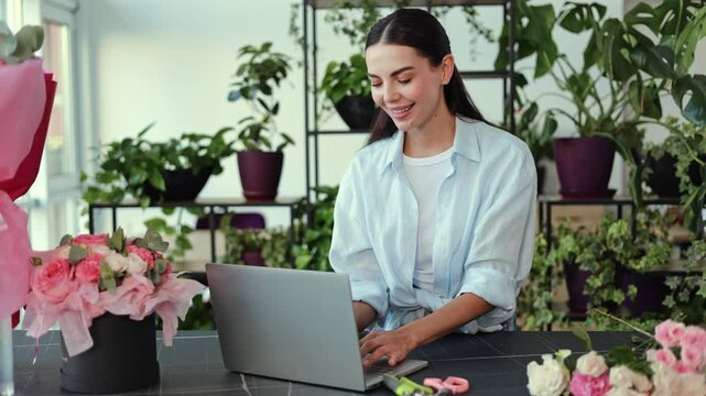 A happy florist smiles at the camera as she stands in her flower shop, arranging flowers and typing on her laptop. Beautiful pink and white flowers and lush green plants surround her.