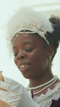 Vertical shot of aristocratic Black woman in feathered fascinator, pearl jewellery and gloves reading letter and smiling outdoors on sunny day