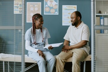 Two professionals engaged in conversation while holding documents in bright medical office setting with posters on wall depicting health advice