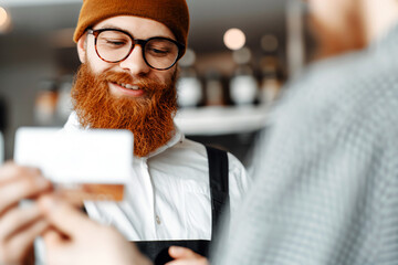 A smiling barista wearing a brown beanie and glasses takes a customer's credit card.
