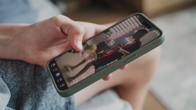 Smartphone application with various video content displayed on phone screen. Close up of teenager scrolling and watching social media feed with short videos using mobile phone while sitting on sofa.