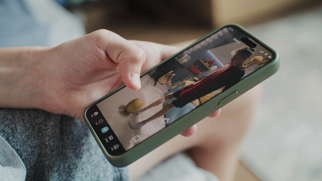 Smartphone application with various video content displayed on phone screen. Close up of teenager scrolling and watching social media feed with short videos using mobile phone while sitting on sofa.