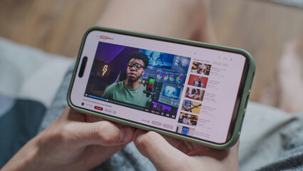 Video sharing platform with blogger content about color correction displayed on smartphone screen. Close up of teenager watching educational video about color grading on mobile phone, sitting on sofa.