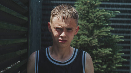 Cinematic shot of Caucasian teen boy with wet face, hair and clothes looking at camera while standing outside in the rain in house yard. Iron fence and coniferous tree in the background. Slow motion. © Framestock