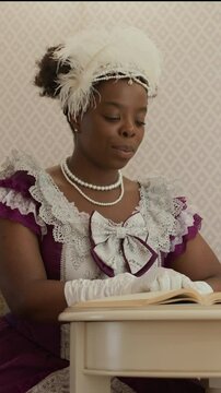 Vertical shot of young Black woman in Victorian-style dress, gloves, pearl necklace and feathered fascinator reading book while sitting at table in living room