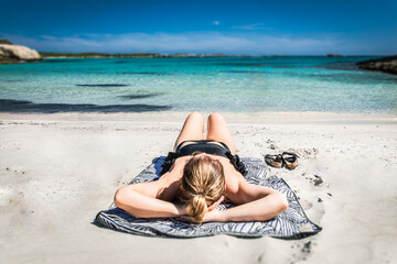 A woman sunbathes on a beach, lying on a towel near the crystal-clear water’s edge. In the background, calm sea and clear sky. A perfect shot of vacation relaxation on a sunny beach.