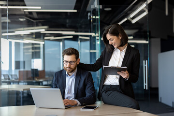 Business colleagues collaborating in modern office. Man in suit working on laptop while woman reviews notes on digital tablet. Teamwork and productivity concept in corporate environment.