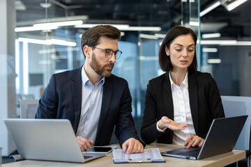 Two colleagues in suits engaged in serious business discussion at office desk. Open laptops and...