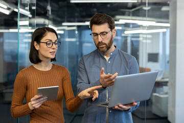 Colleagues engage in discussion in modern office setting. Woman holds tablet, man gestures at...
