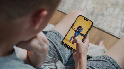 Teen boy scrolls through and watches social network feed with short videos using mobile phone while sitting on sofa. Smartphone app with various video content displayed on phone screen. Close Up.