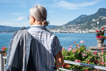 Fototapeta premium Rear view of senior gray haired woman standing along lake admiring the landscape - relaxed elderly grandmother enjoying peaceful moments in vacation, Lake Geneva and mountains in background