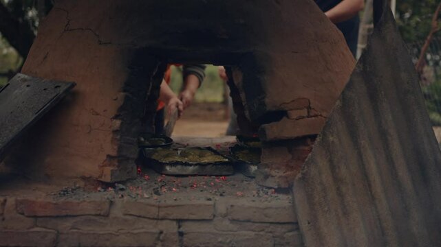 Woman placing Paraguayan chipa into the tatakua, a traditional handmade clay oven from Paraguay. The fire is strong, and smoke is rising from the ground