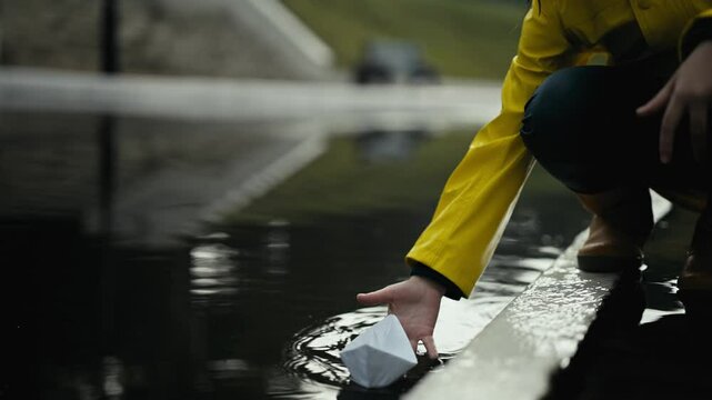 Close-up a teenage girl in a yellow jacket launches a white paper boat into the water through a large puddle during the rain on the street