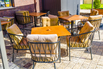 The charming outdoor seating area features plush cushions on steel chairs and lovely wooden tables in a bright, sunlit ambiance, no people