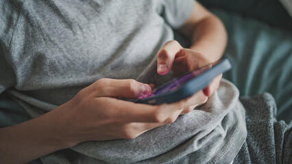 Close up of Caucasian teenager typing and chatting with friends in messaging app using mobile phone, sitting on sofa in living room. Young boy resting and spending weekend at home. Lifestyle concept.