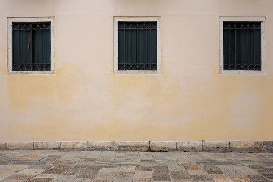 A pale yellow wall with three barred windows and a weathered stone pavement. Perfect for minimalistic design, architectural backgrounds, or projects with a focus on urban simplicity.