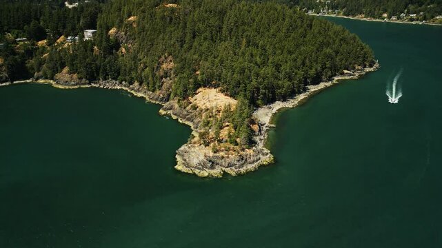 Drone shot of speed boat sailing around a rocky peninsula on Bowen Island in Vancouver with hilly forests and surrounded by tropical blue ocean water on a sunny summer day